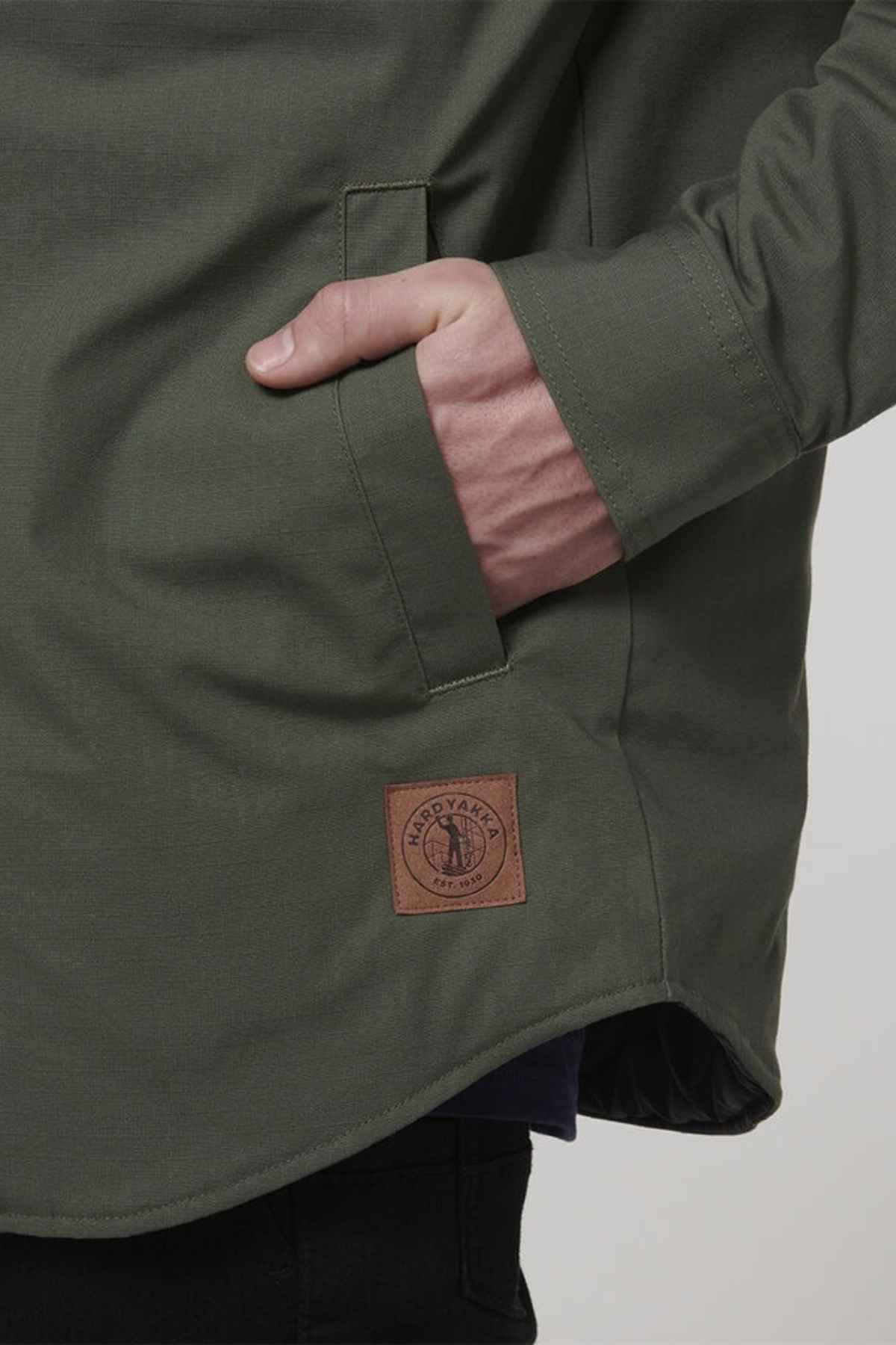 A person wearing the Hard Yakka Quilted Solid Shacket in green, made from durable ripstop fabric, with their right hand in the pocket. The shacket features a brown leather patch with the emblem and text "Handyman" on it. The background is plain and light-colored.