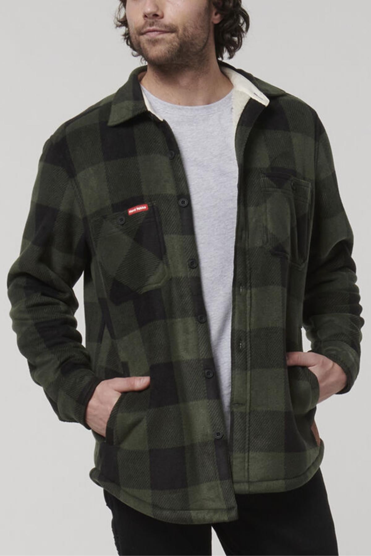 A man is wearing a green and black plaid Hard Yakka Sherpa Bonded Fleece Jacket with a gray T-shirt underneath. He has his hands in the jacket pockets and stands against a plain gray background. The comfortable jacket also features a chest pocket on the left side and resembles a classic Hard Yakka design.