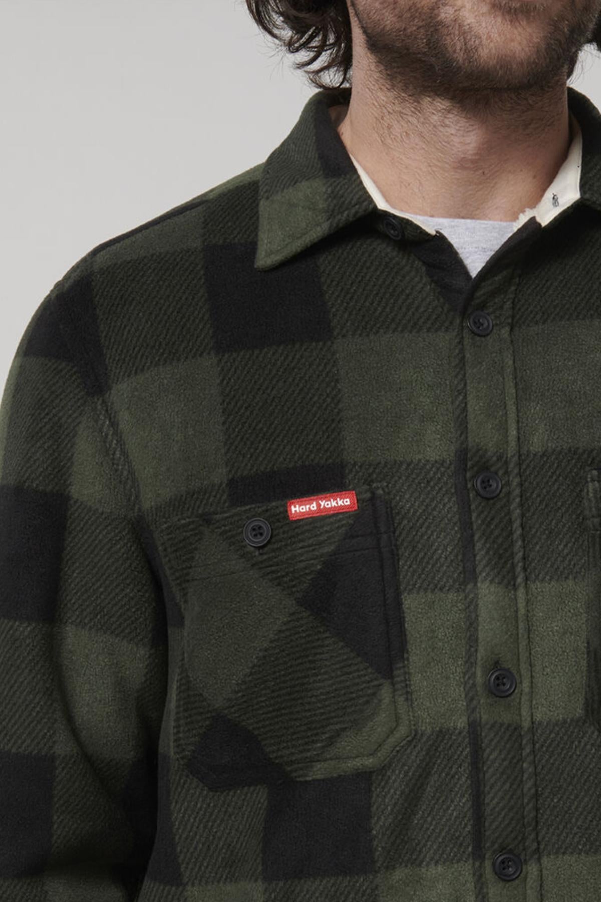 A person wearing a Hard Yakka Sherpa Bonded Fleece Jacket in green and black plaid is shown in the image. The upper torso and part of the face are visible, showcasing the jacket's texture and pattern. A red label on the chest pocket reads "Hard Yakka," reflecting both style and comfort.