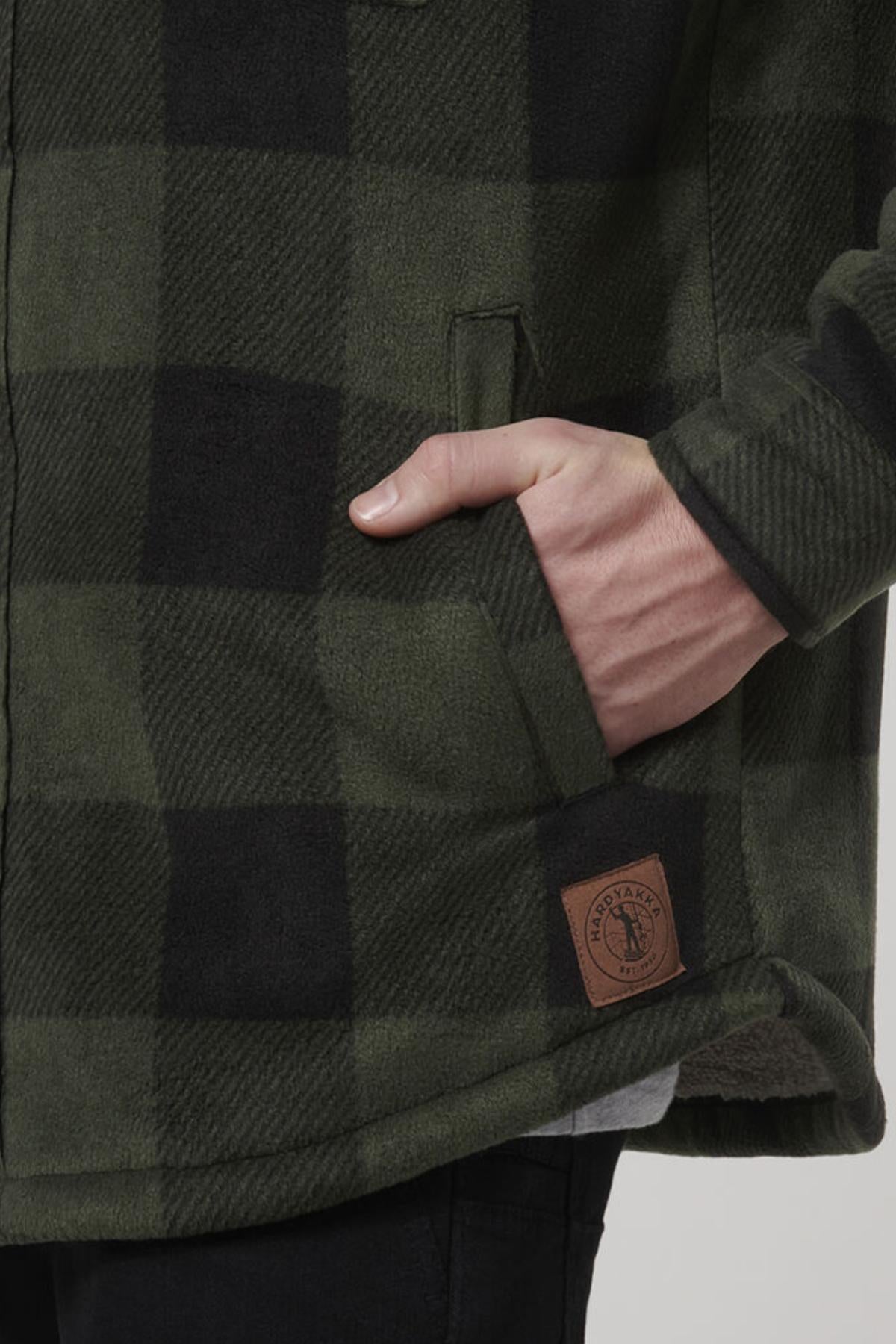 A person wearing a green and black checkered Hard Yakka Sherpa Bonded Fleece Jacket with one hand in the pocket. The comfortable jacket features a small brown rectangular patch with an insignia and the words "HAND CRAFTED" visible on the lower front. The person is also wearing black pants.