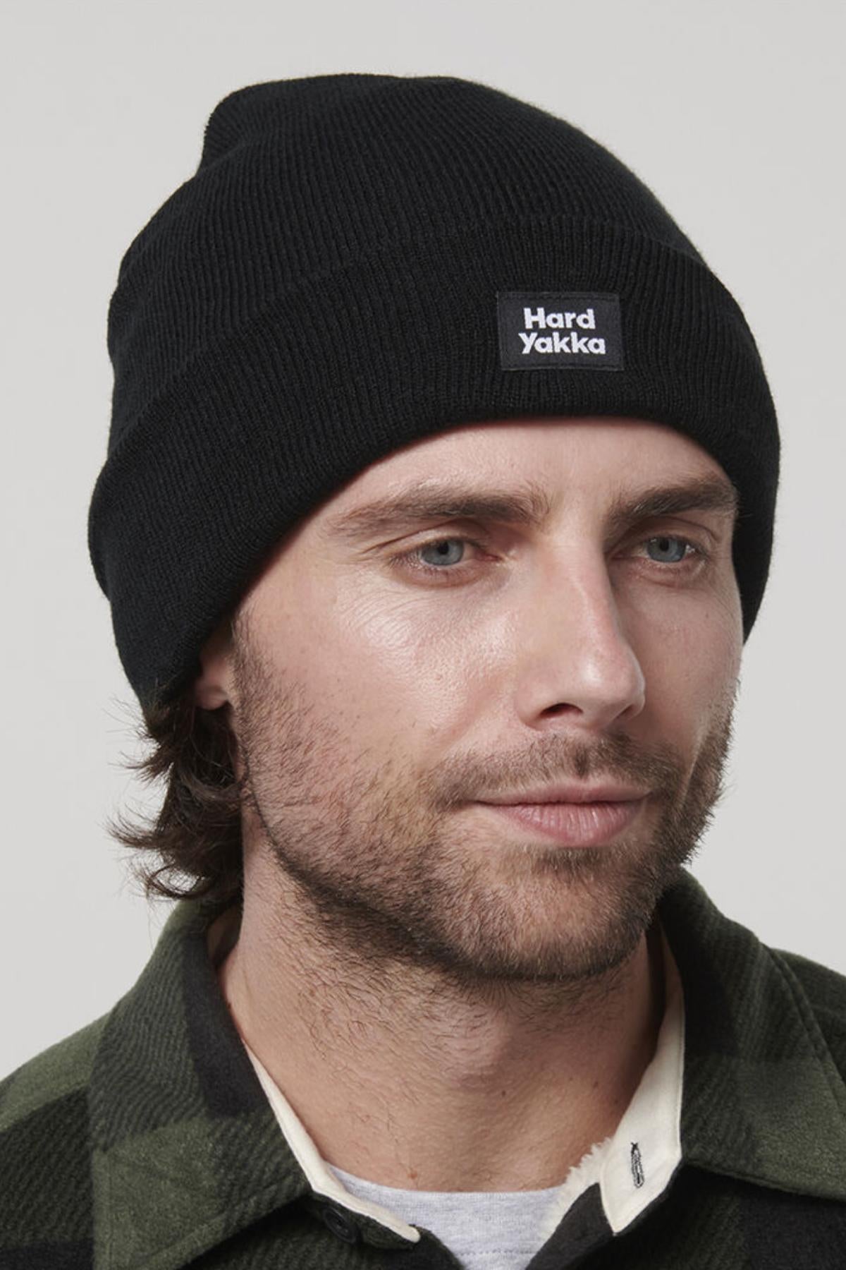 A man with a light beard and blue eyes wears a black beanie featuring the "Hard Yakka" label on the front. He is also sporting a green and black plaid Hard Yakka Sherpa Bonded Fleece Jacket, which suggests the comfort of sherpa lining. His neutral expression gazes slightly past the camera, set against a plain, light-colored background.