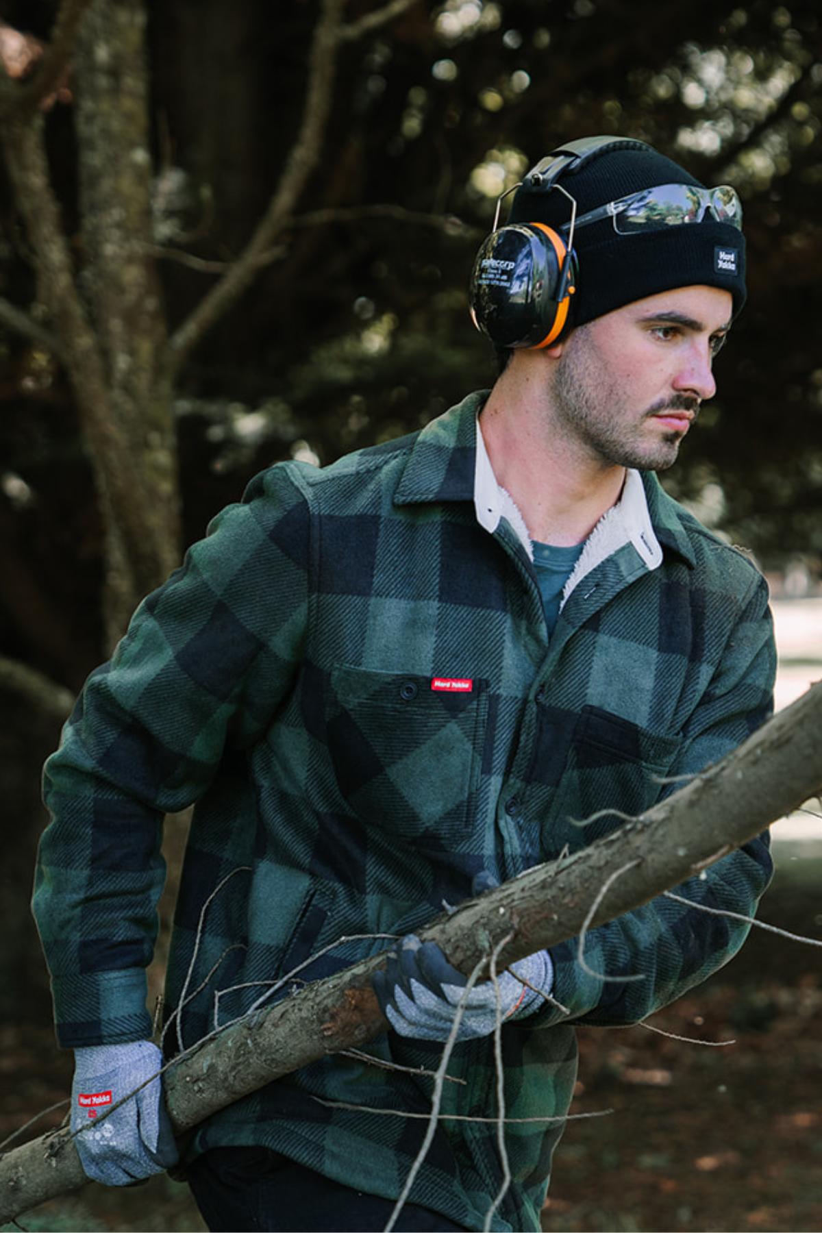 A man wearing a green plaid shirt, gray gloves, a Hard Yakka Sherpa Bonded Fleece Jacket, a black beanie, and safety earmuffs is carrying a large tree branch. The background features trees and foliage.