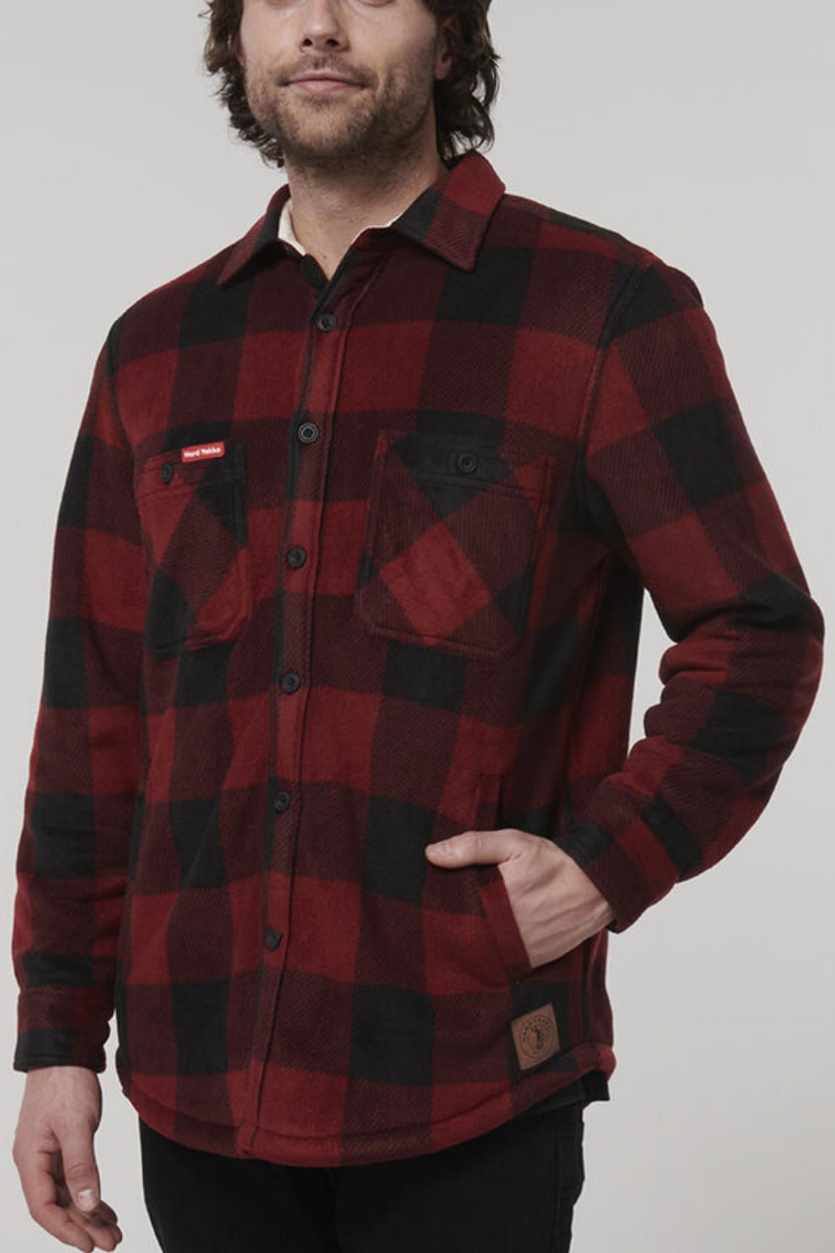 A person wearing a Hard Yakka Sherpa Bonded Fleece Jacket in red and black plaid, which features adjustable cuffs, two chest pockets, and a small brown patch near the hem. The jacket's cozy sherpa lining provides added warmth. They are paired with dark pants and have one hand casually in their pocket.