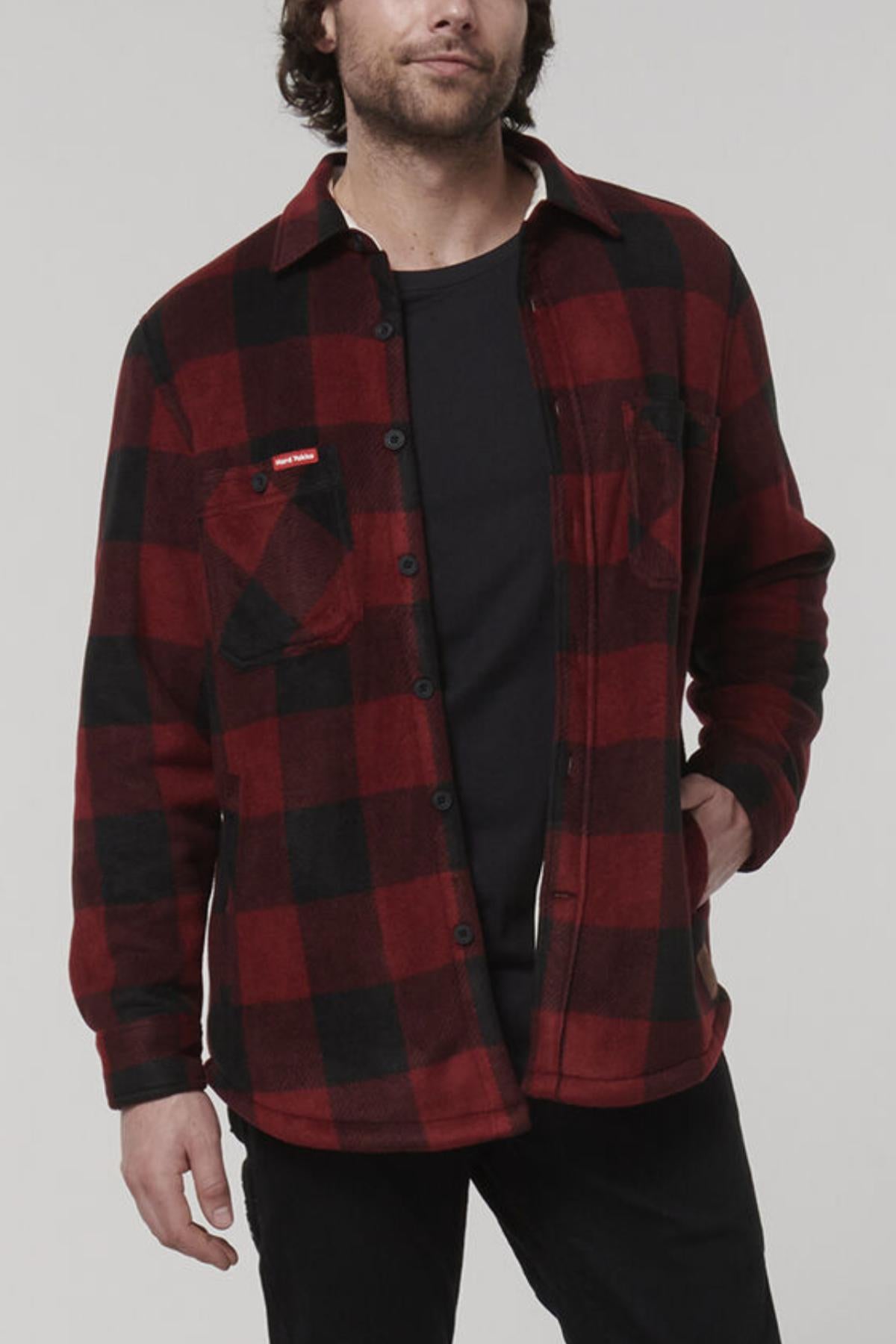 A person wearing a red and black plaid Hard Yakka Sherpa Bonded Fleece Jacket over a black t-shirt. The jacket features two chest pockets, one adorned with a red tag. They have their left hand in their pants pocket and are standing against a plain background.