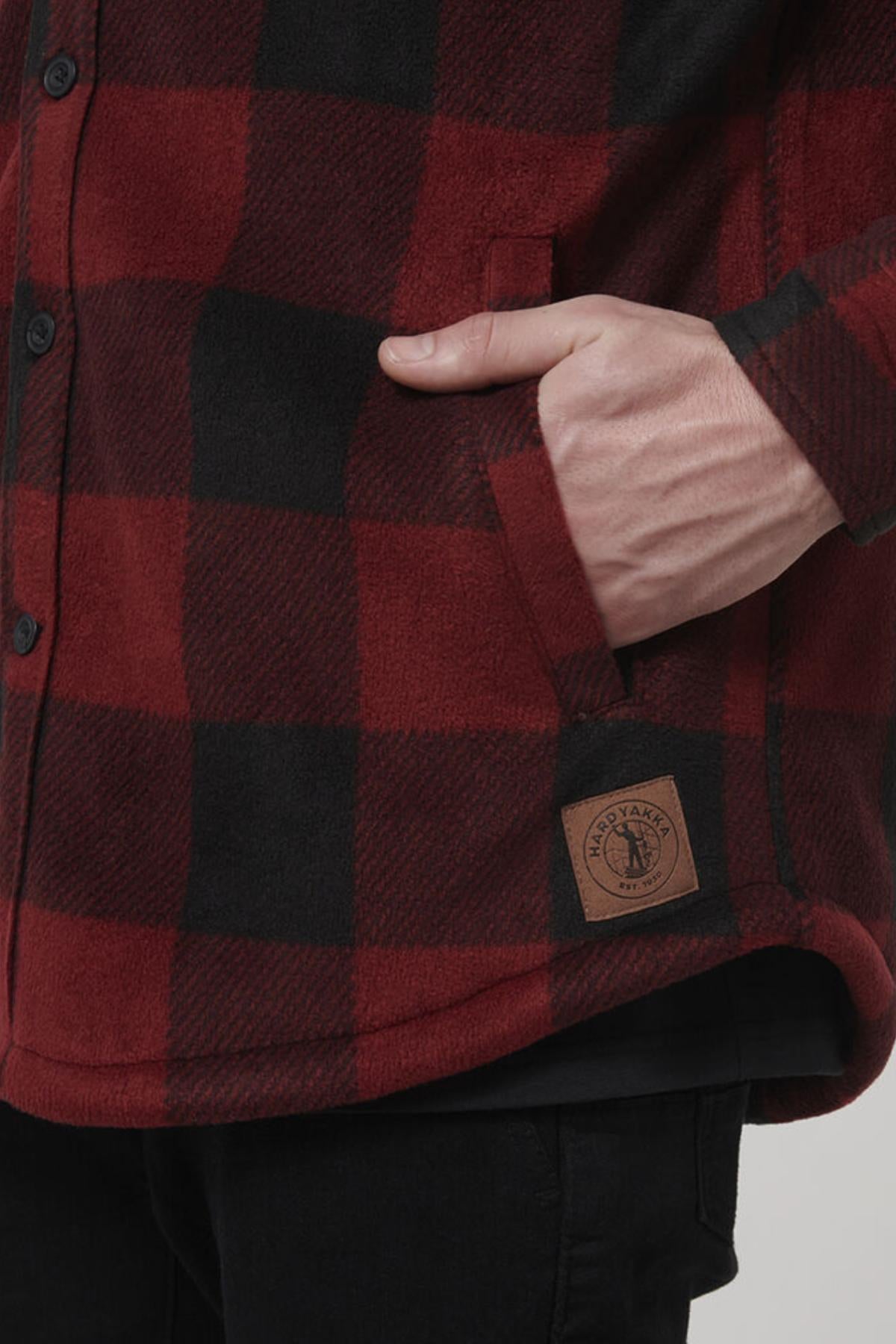 A person with a hand in their pocket models the Hard Yakka Sherpa Bonded Fleece Jacket, featuring a red and black checkered pattern. The jacket is designed with black buttons, adjustable cuffs, sherpa lining, and a brown stitched label near the hem. The person completes the look with black pants.