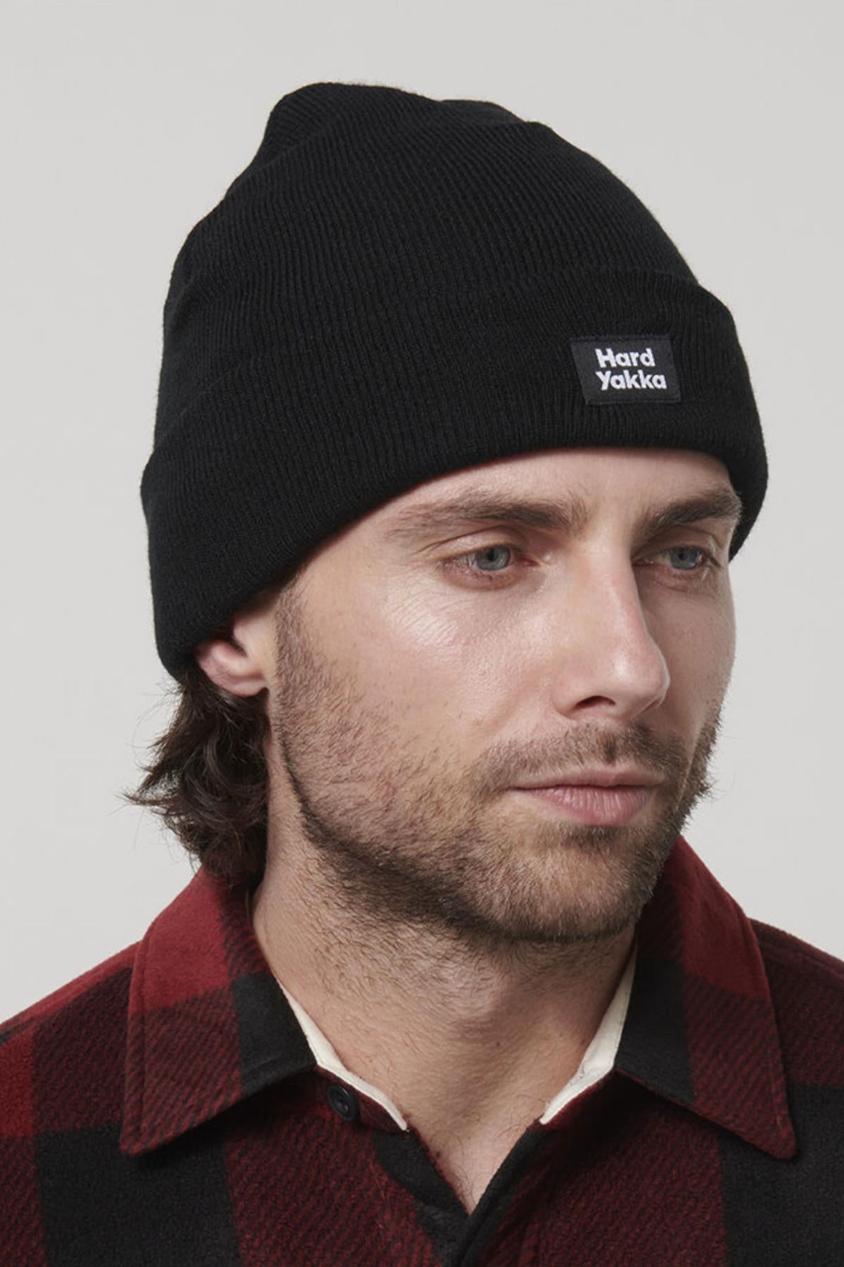 A man with medium-length hair and a beard is wearing a black knitted beanie with a "Hard Yakka" logo on the front. He is also sporting a Hard Yakka Sherpa Bonded Fleece Jacket, designed in red and black plaid. He gazes slightly to the side, and his jacket features adjustable cuffs. The background is plain and light-colored.