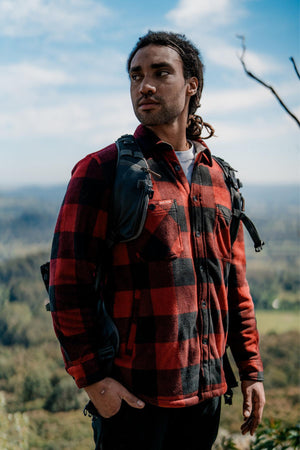 A person with long hair wears a red and black Hard Yakka Sherpa Bonded Fleece Jacket and carries a black backpack, standing outdoors with a scenic view of mountains and forests in the background. The individual appears thoughtful and is looking slightly to the side.