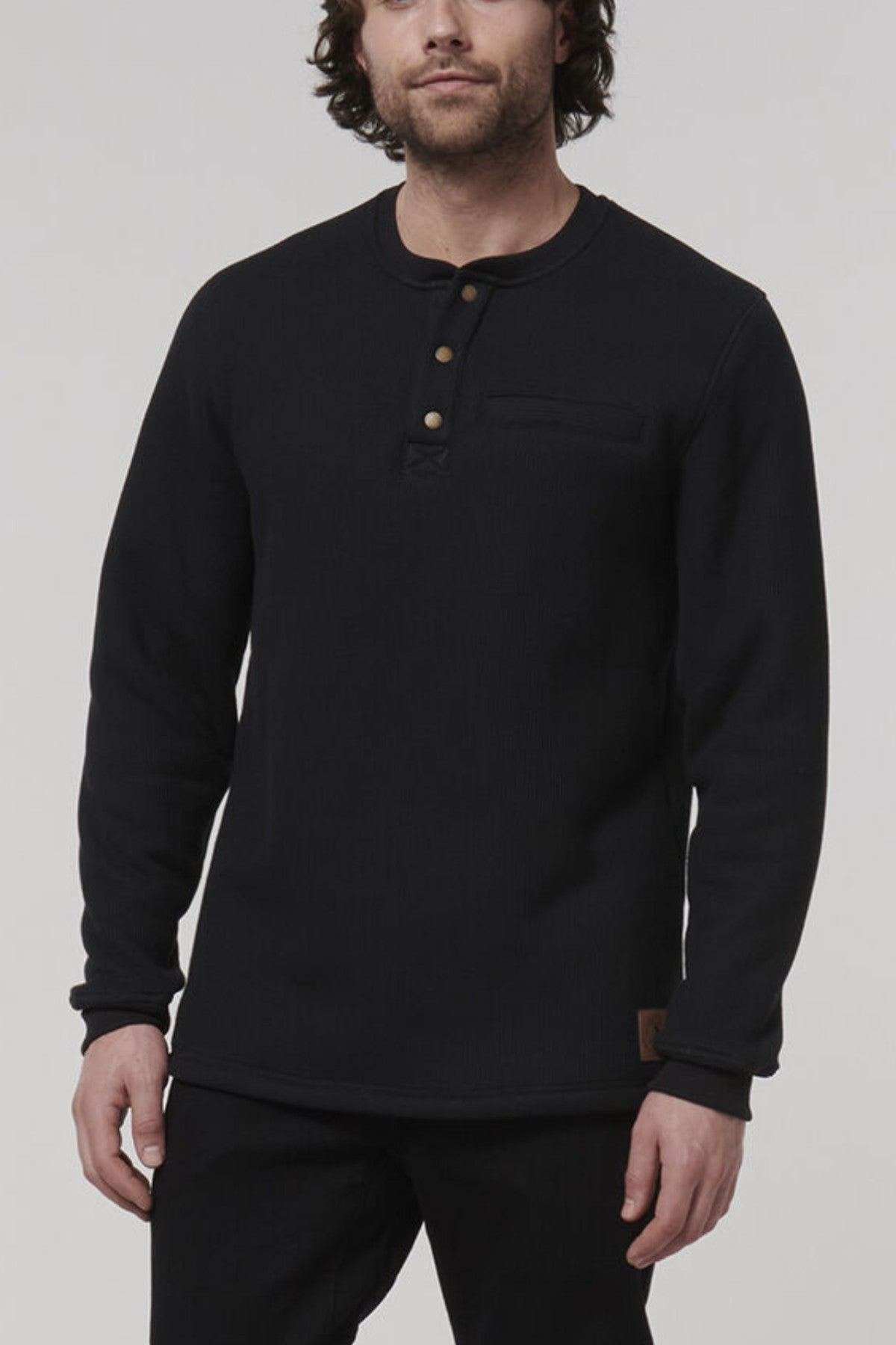 A man models the Hard Yakka Sherpa Henley, a regular fit black long-sleeve shirt with a buttoned collar. It has a small patch near the hem, and he stands against a neutral backdrop with his hands relaxed at his sides.
