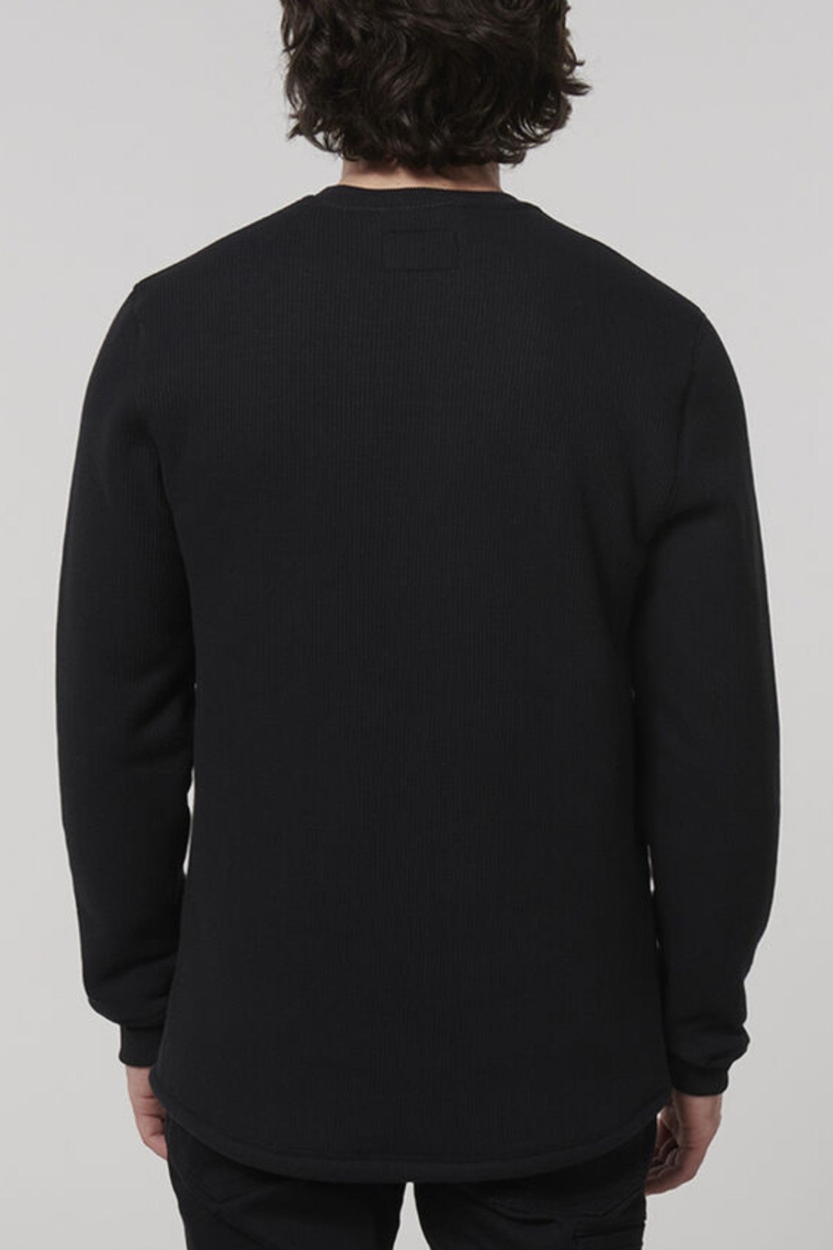 A person with dark wavy hair wears a long-sleeve black Hard Yakka Sherpa Henley, standing against a plain gray background.