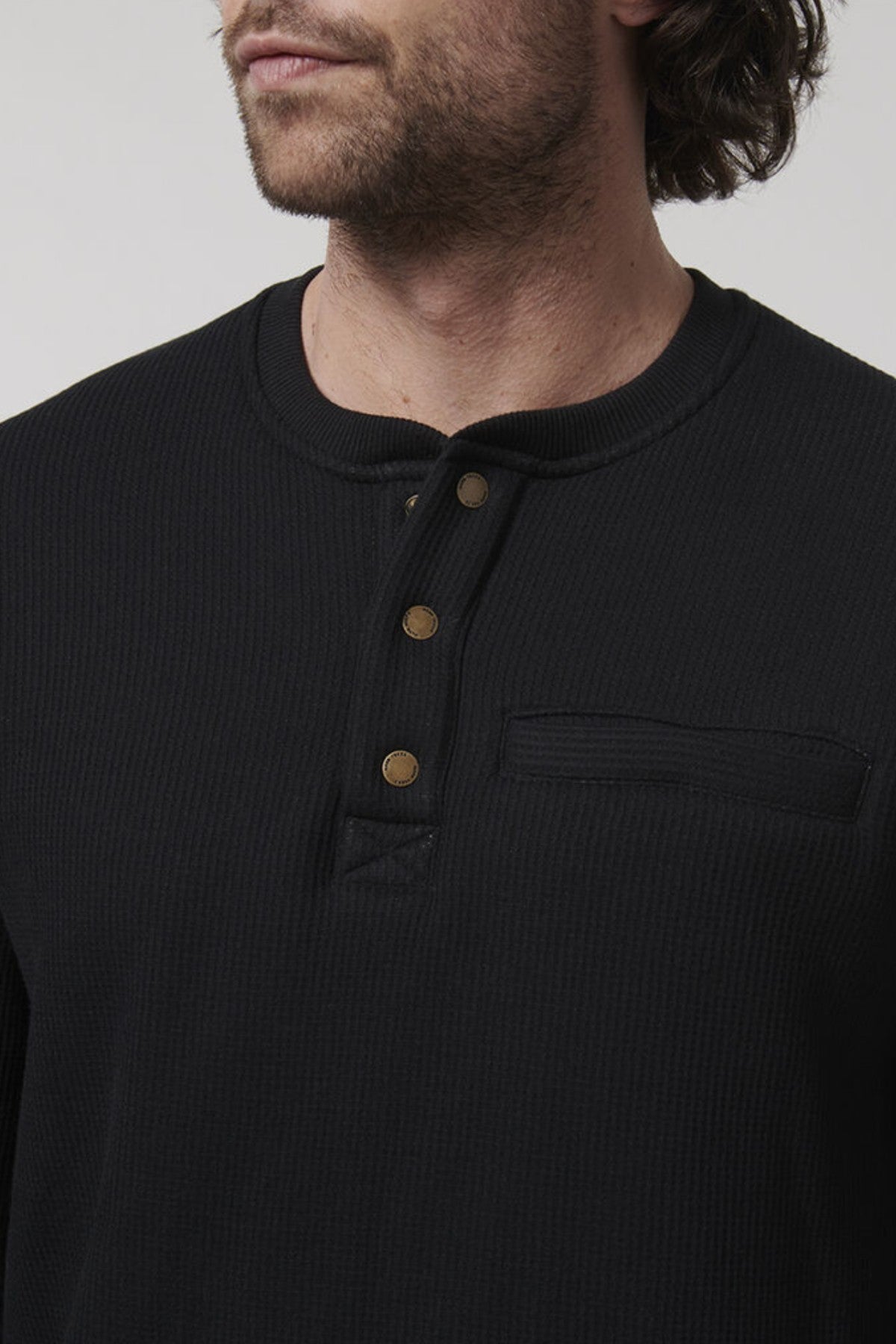 A man with medium-length hair wears a Hard Yakka Sherpa Henley—a regular fit, black shirt featuring three visible brass buttons and a small chest pocket. The image highlights his upper torso and part of his face with a neutral expression.