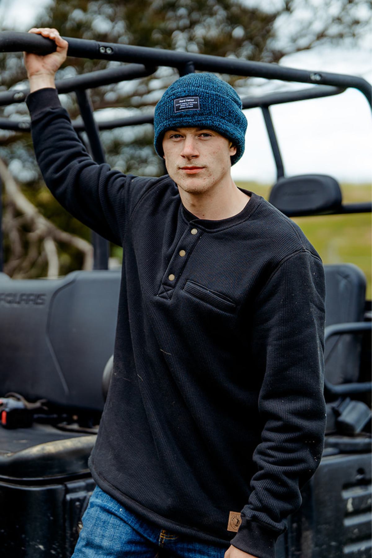 Wearing a blue knit beanie and a Hard Yakka Sherpa Henley, a person leans against an off-road vehicle in an outdoor setting with grassy terrain and trees.