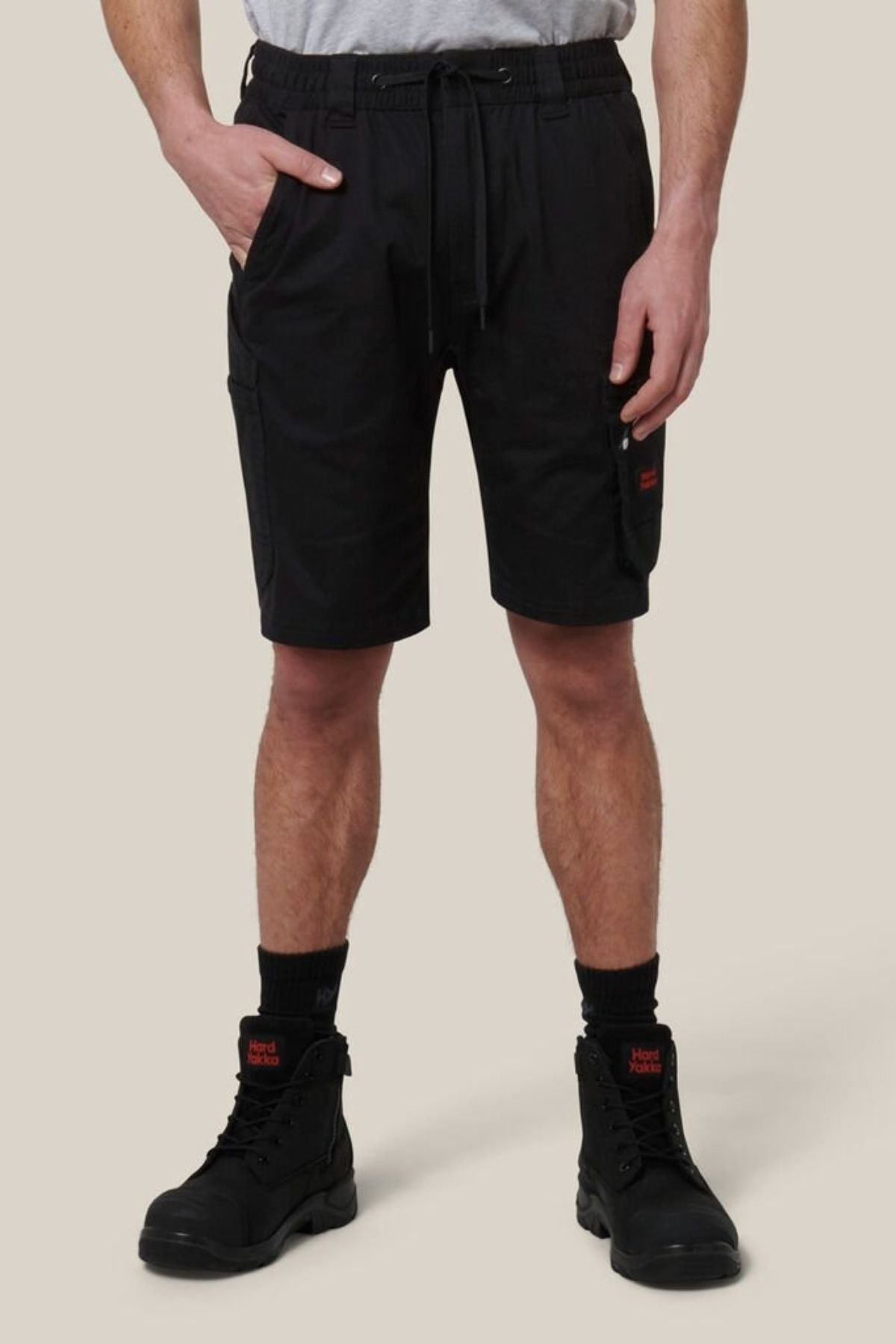 A person wearing a white t-shirt, Hard Yakka Tough Maxx Mid Short with an elastic waistband and drawstring, black socks, and black lace-up boots stands against a plain light beige background.
