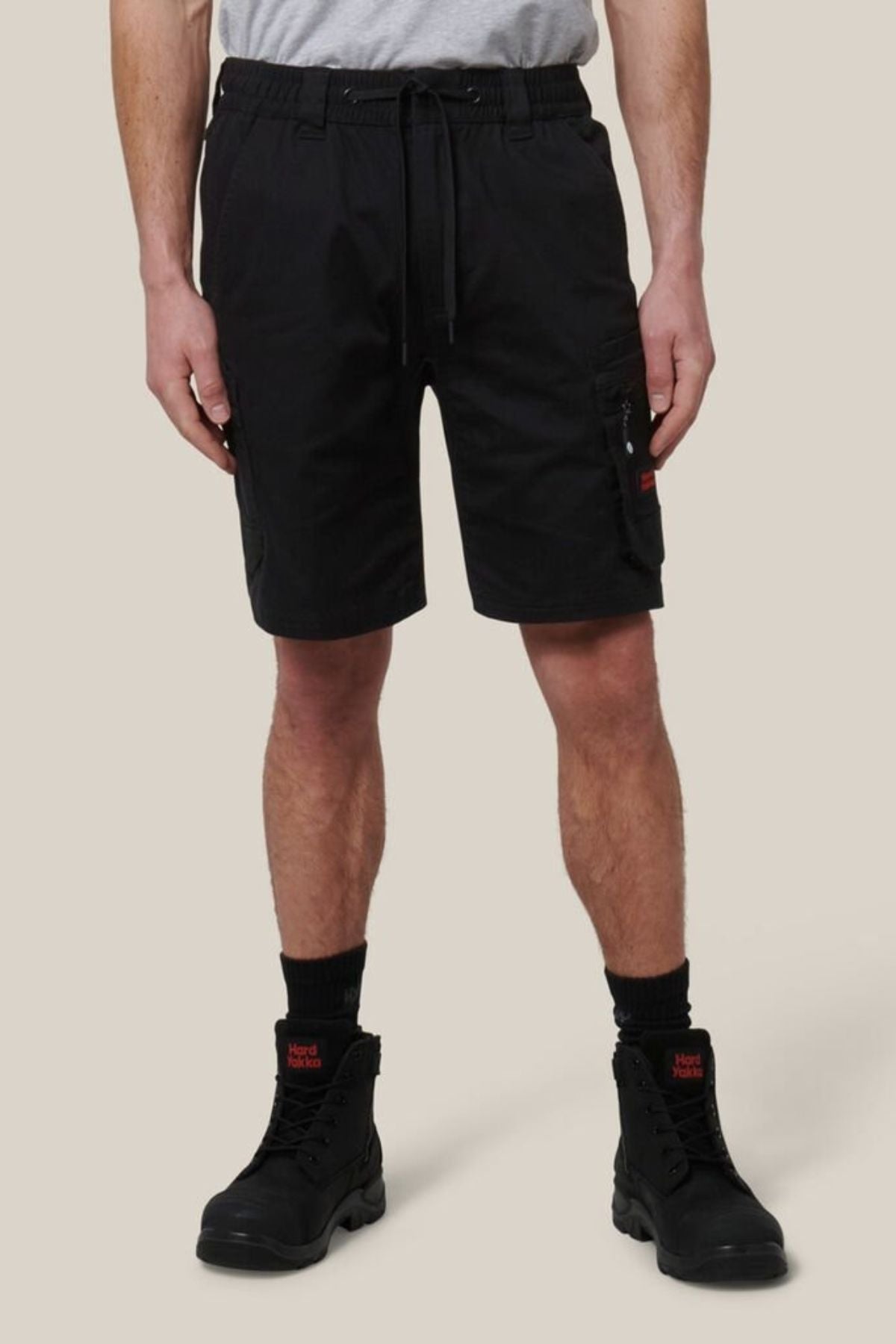 A person in a light gray T-shirt and Hard Yakka Tough Maxx Mid Short with pockets, plus black socks and lace-up boots, stands against a beige background. The shorts provide UPF 50+ protection for extra durability.