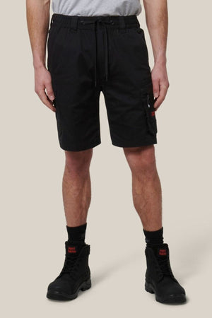 A person in a light gray T-shirt and Hard Yakka Tough Maxx Mid Short with pockets, plus black socks and lace-up boots, stands against a beige background. The shorts provide UPF 50+ protection for extra durability.