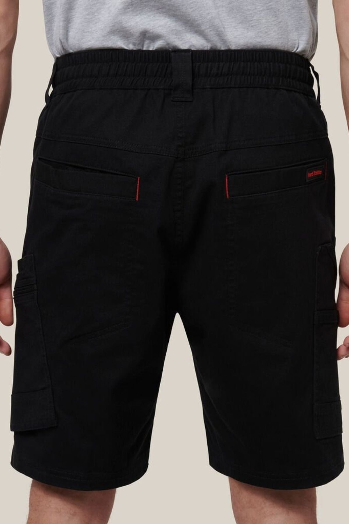 A person wears the Hard Yakka Tough Maxx Mid Short work shorts in black with UPF 50+ protection and red stitching, paired with a gray T-shirt. The back view highlights multiple pockets on the shorts.