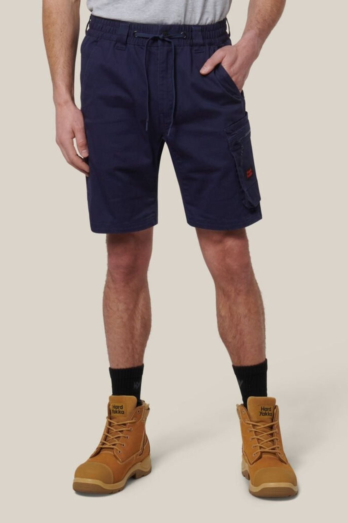 A person wearing Hard Yakka Tough Maxx Mid Short in navy with a matching belt, black socks, and tan work boots stands against a plain light background, showcasing only the lower body.
