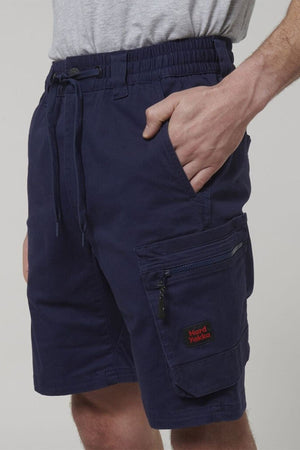 A person wears Hard Yakka Tough Maxx Mid Shorts in grey, featuring UPF 50+ protection, a drawstring waist, zippered side pocket, and a red "Hard Yakka" logo. One hand is tucked into the pocket.