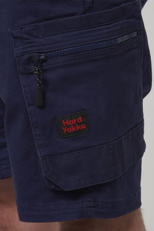 Close-up of a person wearing Hard Yakka Tough Maxx Mid Short work shorts in dark blue, showing a zippered pocket with UPF 50+ protection and a black and red "Hard Yakka" label on the pocket flap.