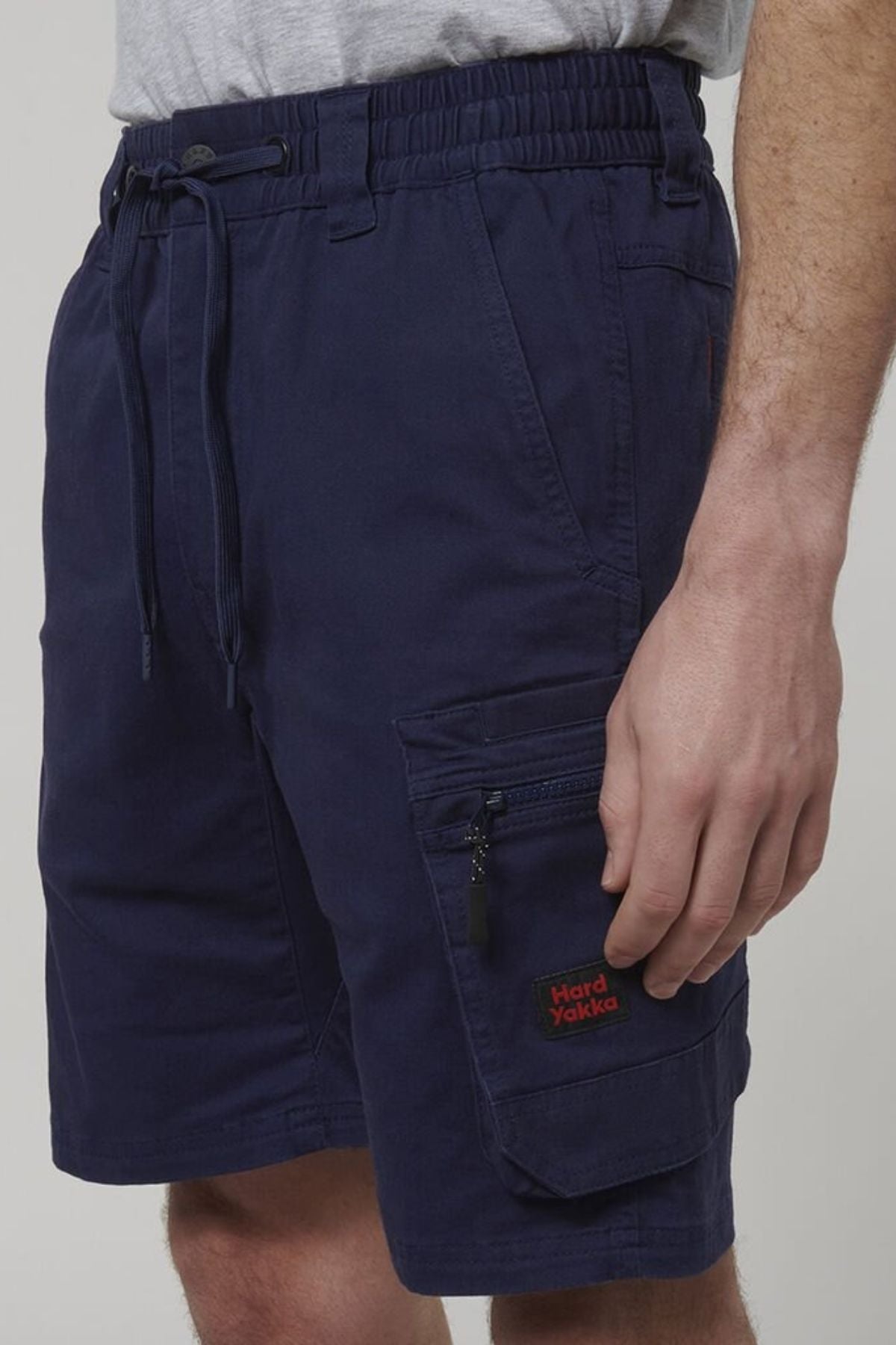 A person wears Hard Yakka Tough Maxx Mid Short work shorts in navy blue, featuring a drawstring waist, zippered pocket with red logo, and UPF 50+ protection, paired with a tucked-in light grey t-shirt.