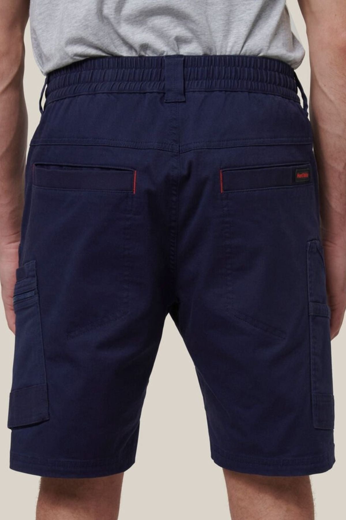 A person wearing Hard Yakka Tough Maxx Mid Shorts in navy, featuring an elastic waistband, two back pockets with red stitching details, and UPF 50+ protection, shown waist down from the back.