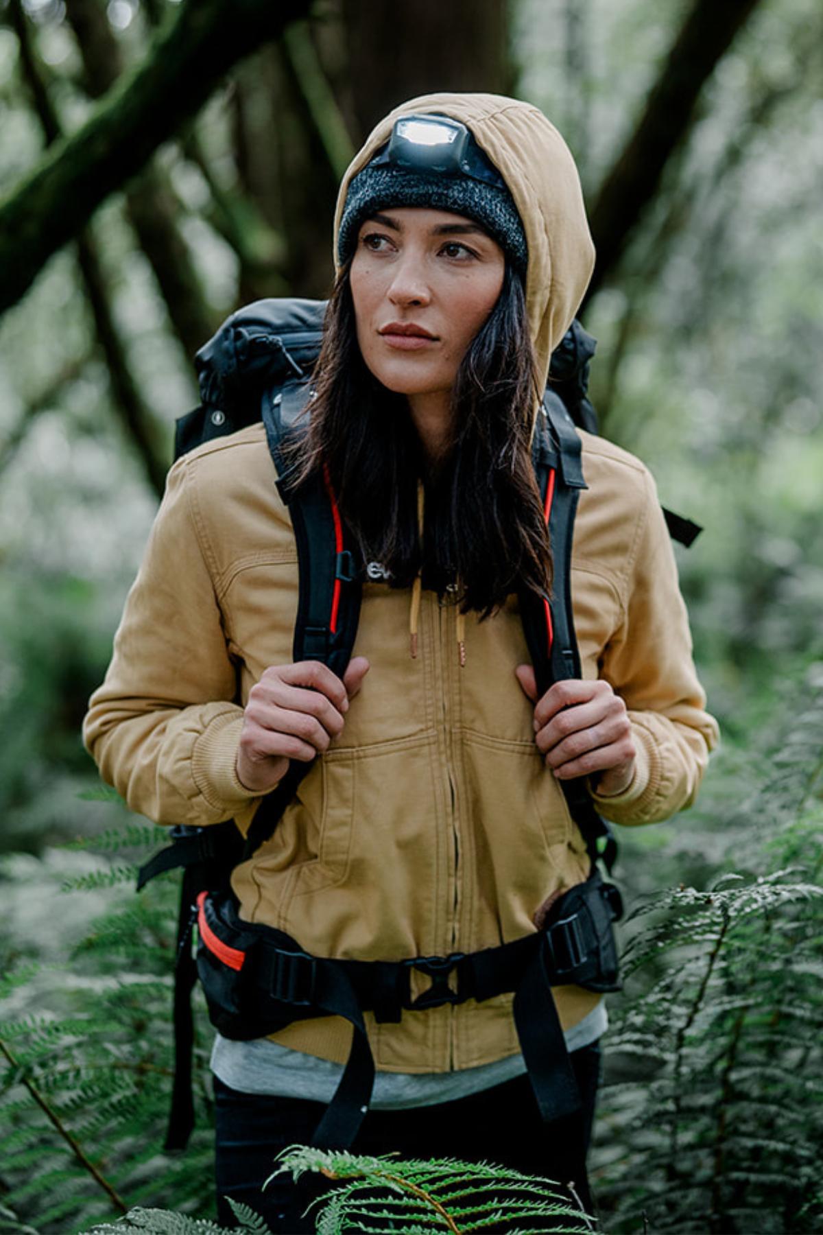 A person in a tan Hard Yakka Women's Bomber Jacket with fleece lining, plus a beanie and headlamp, stands in a forest. With a black backpack, they look to the side amid green foliage and trees.