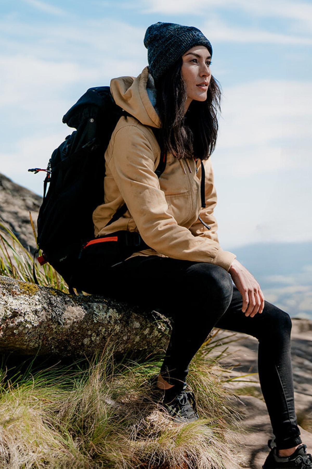A woman sits on a rock outdoors, wearing a Hard Yakka Women's Bomber Jacket with fleece lining, black leggings, a navy beanie, and hiking shoes. She has a large black backpack and gazes into the distance. Grass and a cloudy sky are visible.