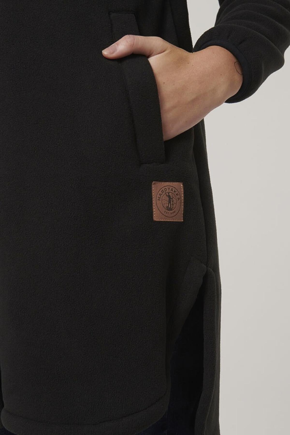 A close-up of a person’s hand in the pocket of a black Hard Yakka Womens Longline Zoodie, featuring a brown rectangular logo patch on the lower side and enhanced thermal insulation for extra warmth.