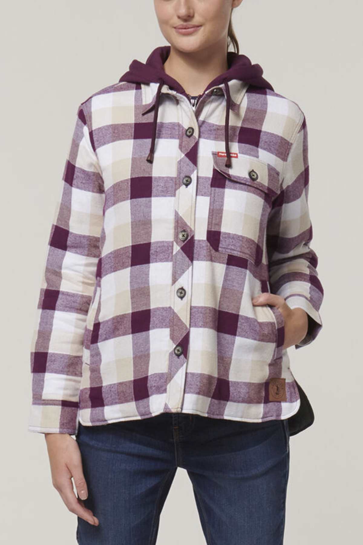 Wearing a Hard Yakka Women's Quilted Shacket in purple and white plaid with button closures, a front pocket, quilted lining for warmth, and a small red tag on the chest pocket, paired with blue jeans. One hand rests in the jacket pocket.