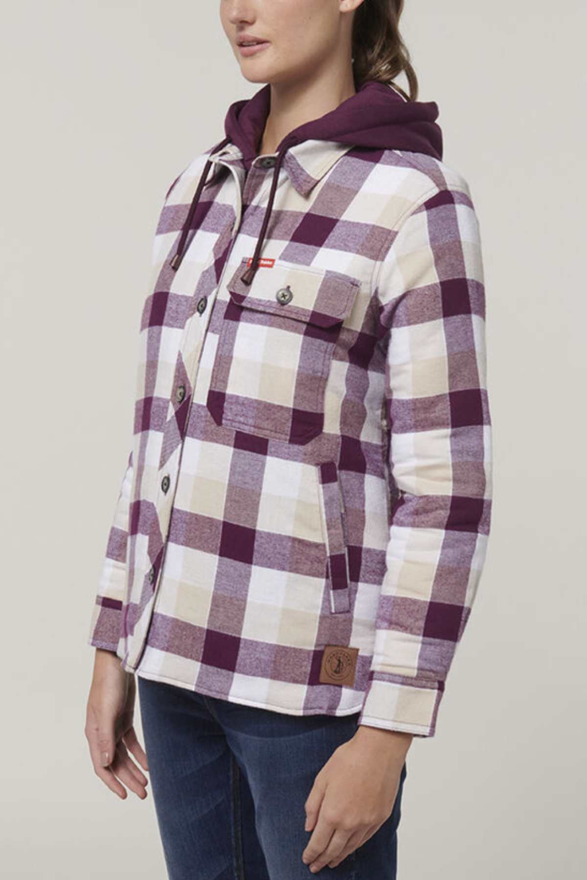 The individual wears a Hard Yakka Women's Quilted Shacket in purple and white plaid with a warm quilted lining and convenient front buttons and a chest pocket, layered over a purple hoodie, paired with dark jeans, all set against a plain background.