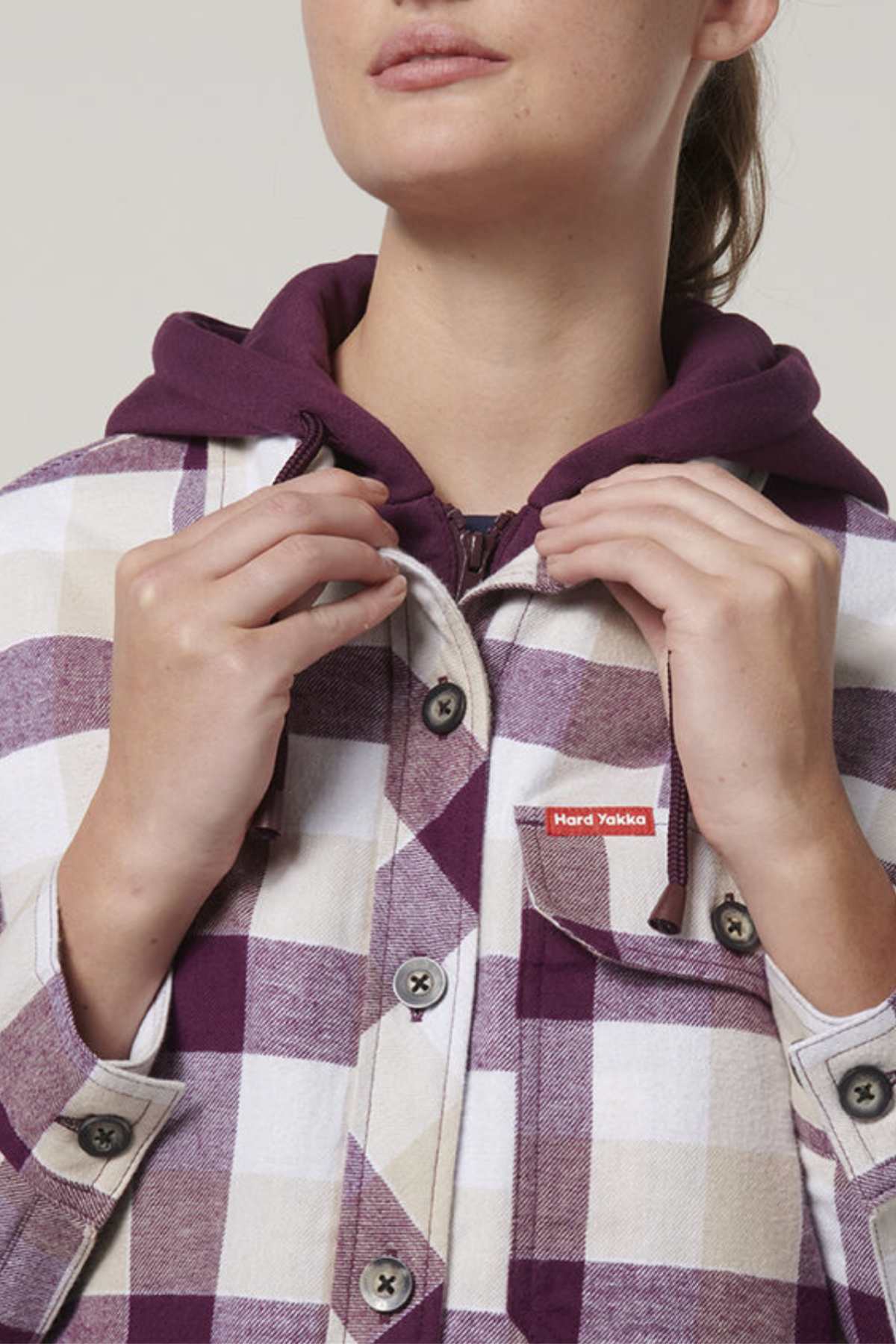 A person wears a purple hoodie under the Hard Yakka Women's Quilted Shacket, showcasing a checkered pattern in purple, beige, and white. They hold the buttons of the shacket, which features a red and white label on its pocket.
