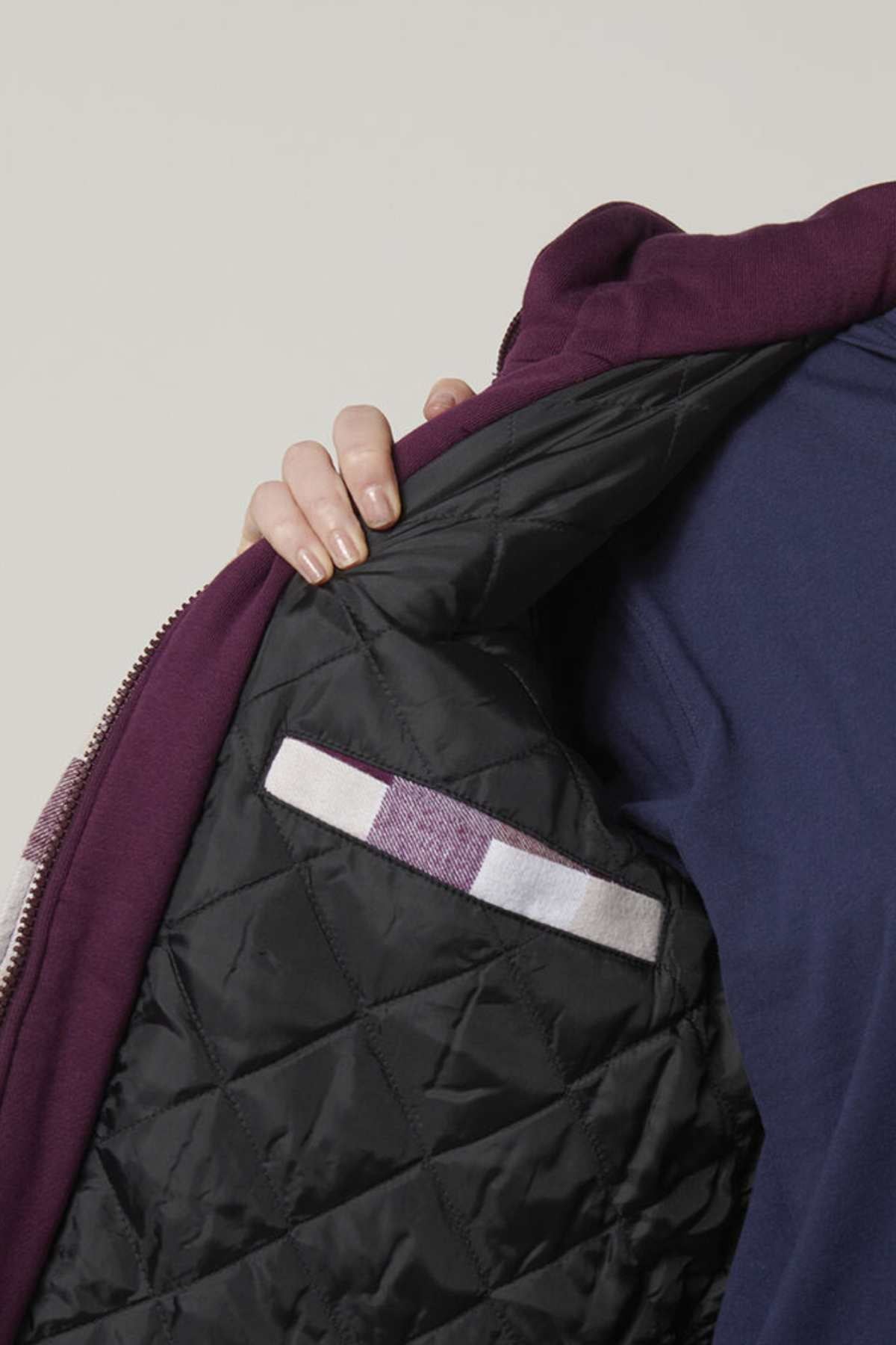 A person showcases the Hard Yakka Women's Quilted Shacket, revealing its black quilted lining and white and purple striped label, perfectly pairing it with their blue shirt.