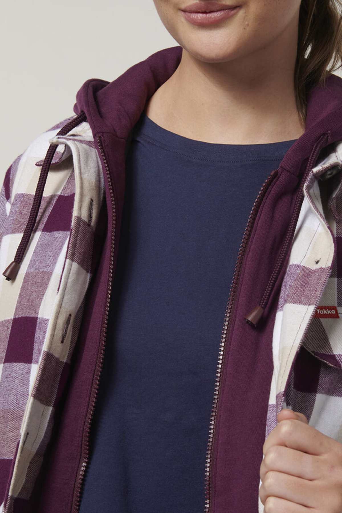 Wearing a "Hard Yakka Womens Quilted Shacket," this person pairs it with a blue shirt and purple hoodie. The partially zipped, purple-and-white checkered jacket has a durable quilted lining, and they grasp its edges with one hand, revealing only part of their face.