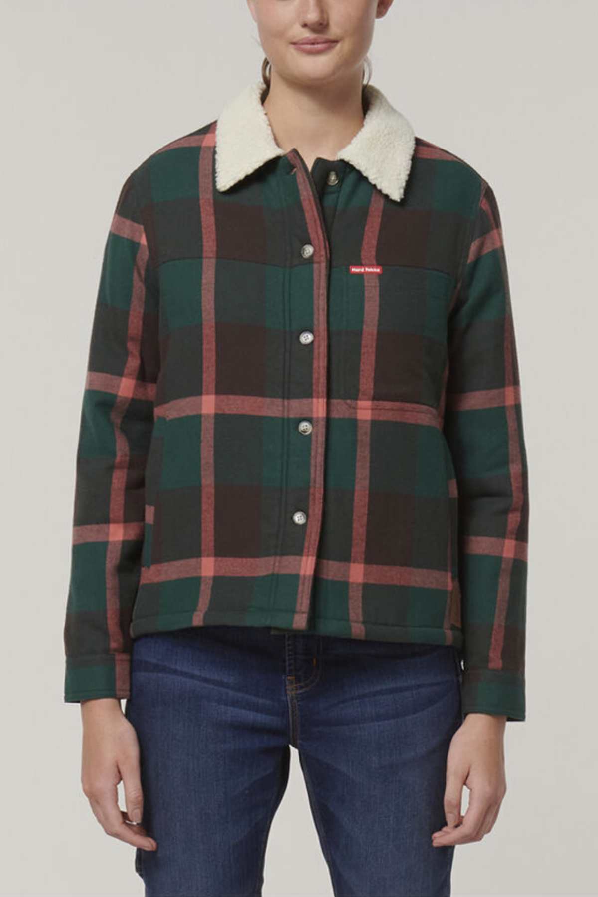 Wearing the Hard Yakka Women's Heritage Trucker Sherpa Jacket in green and pink plaid with a sherpa collar, a person stands against a plain background. The jacket features a chest pocket and front buttons, paired stylishly with blue jeans.