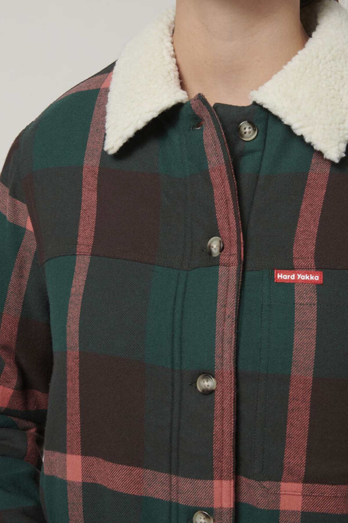 A person models the Hard Yakka Womens Heritage Trucker Sherpa Jacket—a regular fit plaid jacket in green, black, and red featuring a cozy white sherpa collar and "Hard Yakka" chest label. Only the upper torso and neck are visible.
