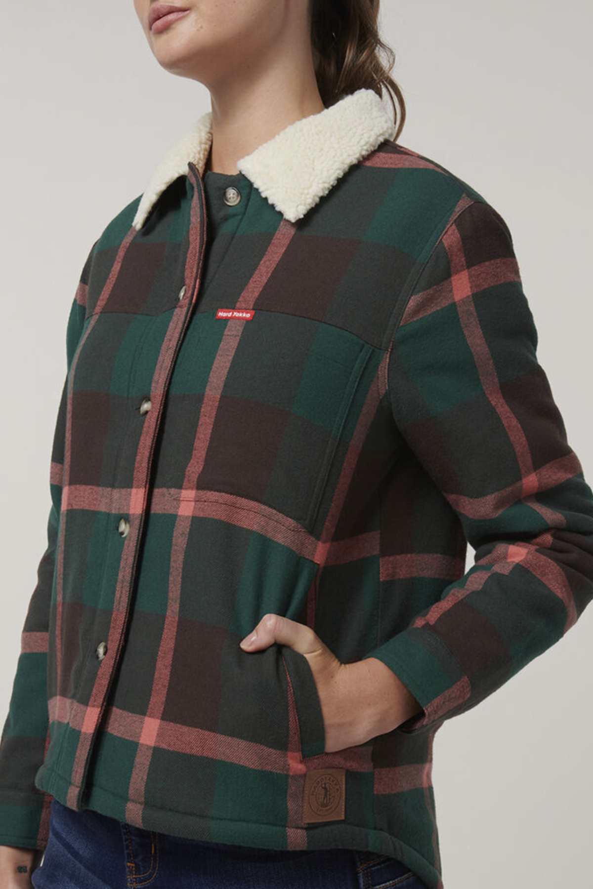 A person wears the Hard Yakka Womens Heritage Trucker Sherpa Jacket, featuring green, brown, and red plaid, a white sherpa collar, snap buttons, a small red chest label, regular fit, with one hand in a pocket.