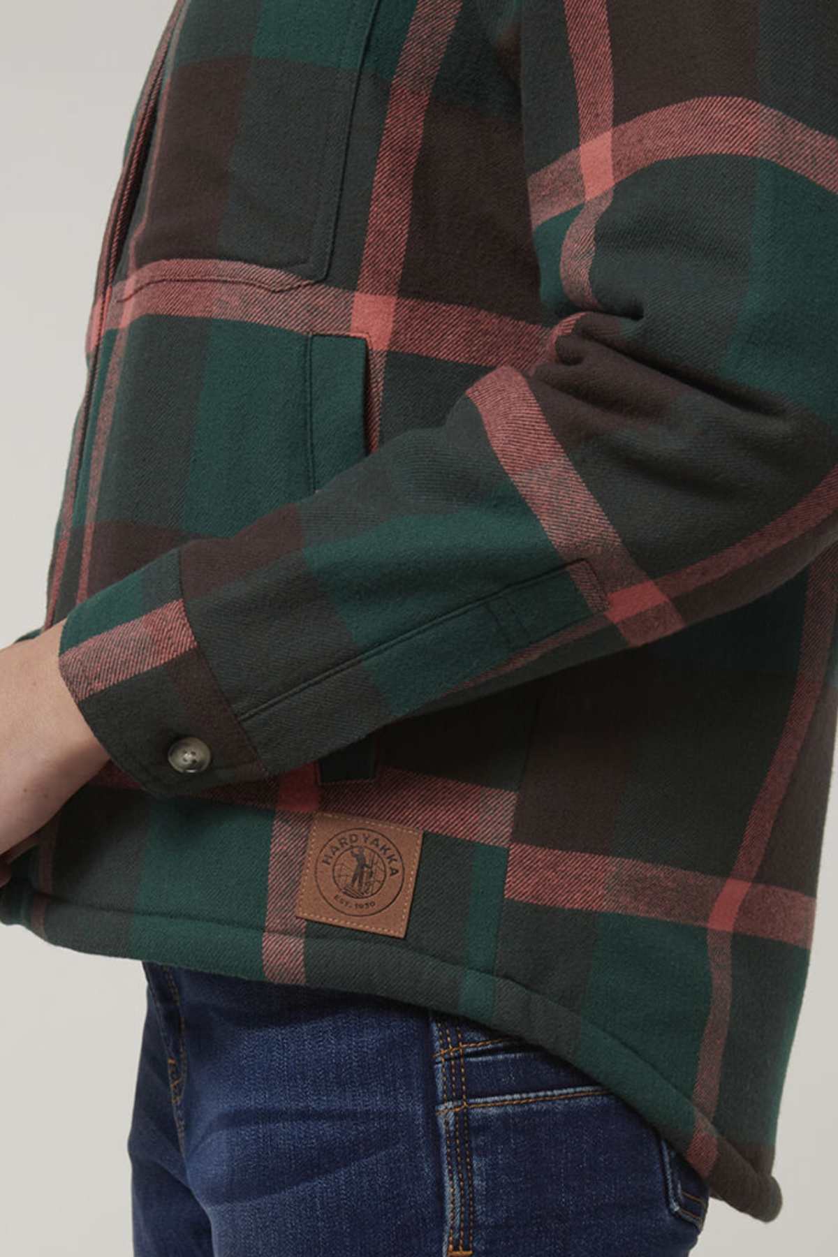 A person wears the Hard Yakka Womens Heritage Trucker Sherpa Jacket in green, brown, and red plaid with a leather patch above the hem, paired with blue jeans. The image highlights the buttoned cuff and side torso details.