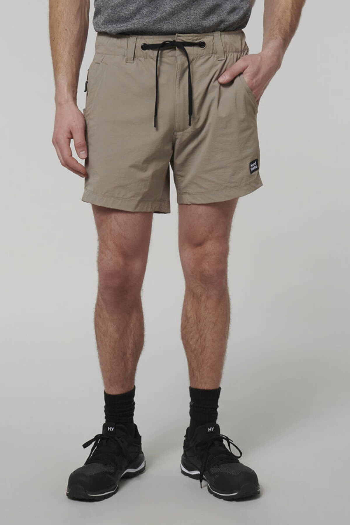 A person in a gray t-shirt and beige Hard Yakka X Short Shorts with a black drawstring has hands in pockets. They're also wearing black socks and sneakers. The shorts, crafted from 4-way stretch Cordura Nylon, pop against the plain light gray backdrop.