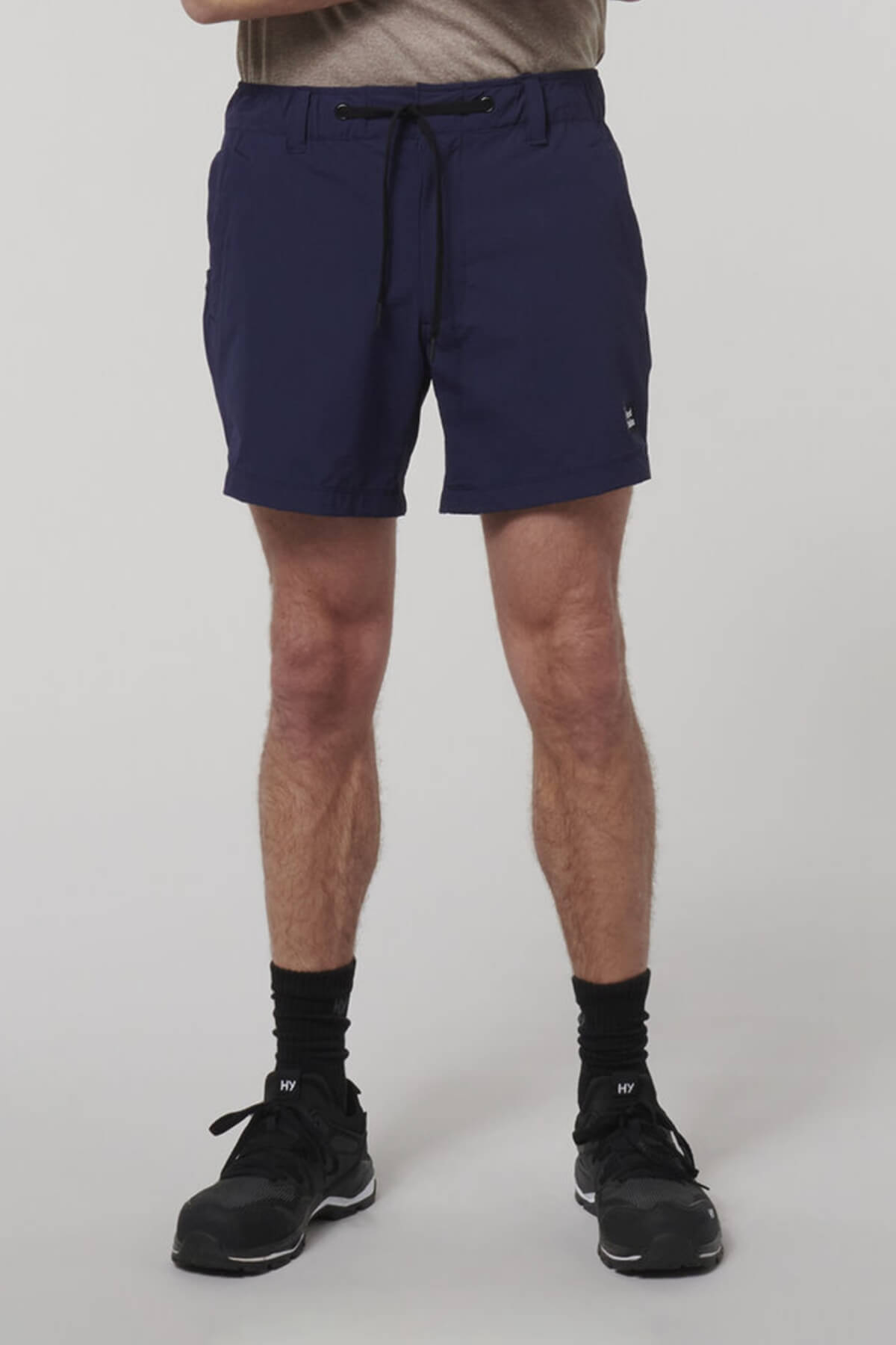 A person models the Hard Yakka X Short Short in navy blue, paired with a tucked-in beige shirt, black socks, and black athletic shoes, standing against a plain light gray background. The image highlights the lower half of the body.