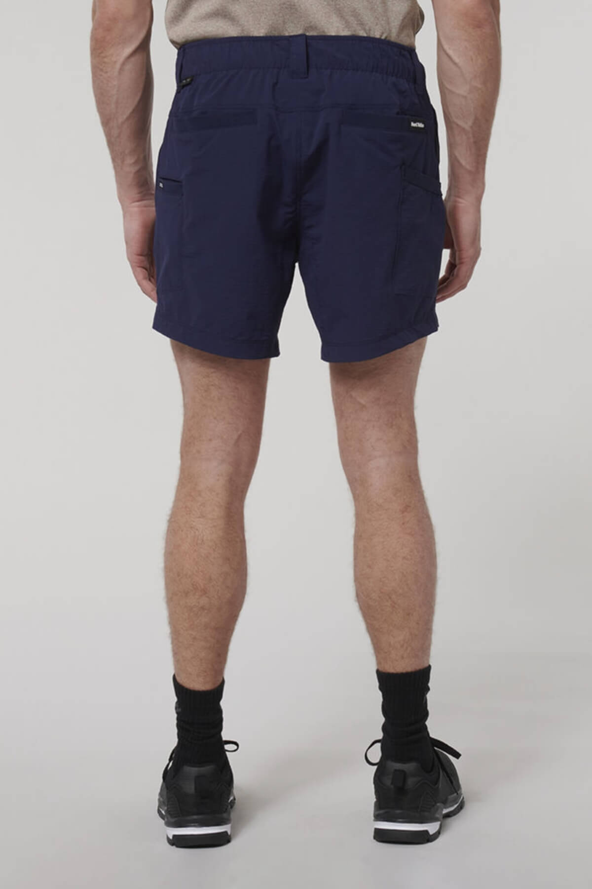 A person stands facing away from the camera wearing the Hard Yakka X Short Short in navy blue, made from 4-way stretch, moisture-wicking fabric, paired with a beige shirt, black socks, and black sneakers against a plain light background.