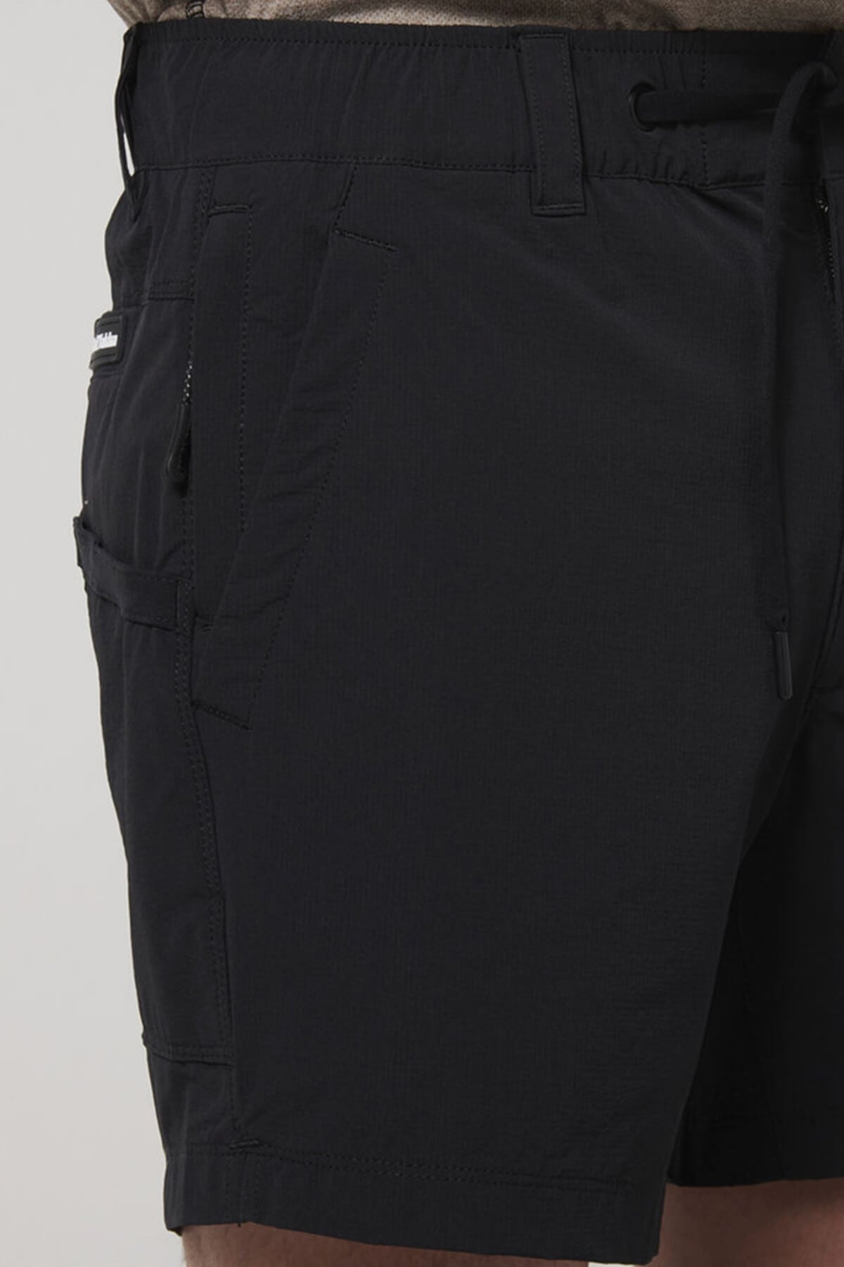 A close-up of a person wearing the Hard Yakka X Short Short in black, featuring an elastic waistband, 4-way stretch fabric, side pockets, and drawstring, shown from waist to mid-thigh against a plain background.