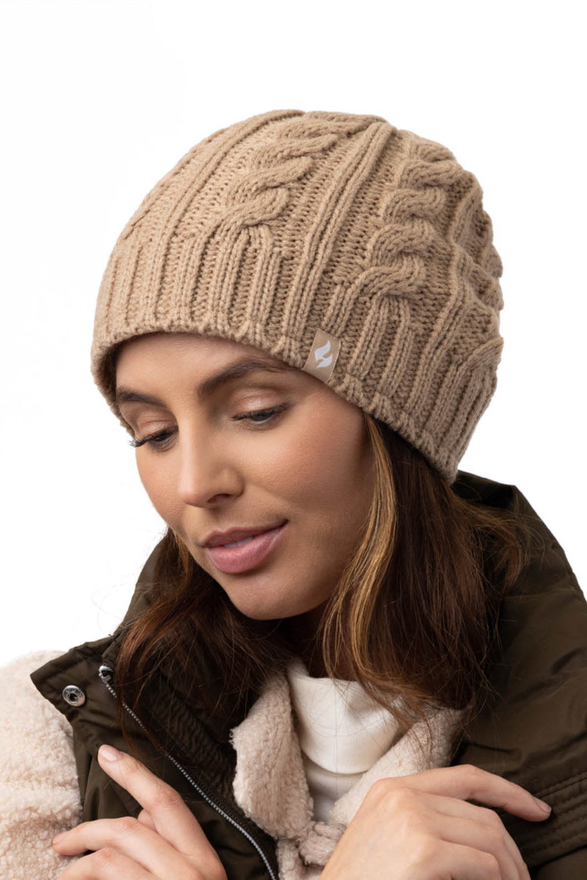 A woman with long brown hair is wearing a beige Heat Holders Alesund Ladies Thermal Beanie and a brown quilted jacket over a white top. She is looking downwards with a soft smile, and her hand is adjusting her Heat Holders jacket collar. The background is plain white.