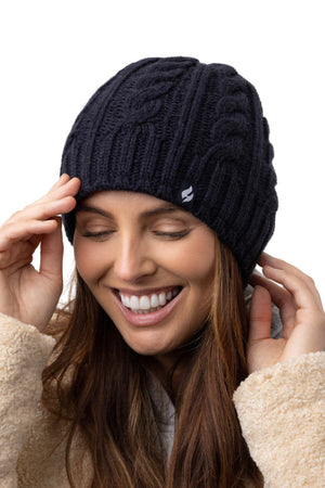 A person with long brown hair smiles warmly while wearing a thick, dark blue Heat Holders Alesund Ladies Thermal Beanie with a classic cable knit design and a beige, fluffy jacket. The person's eyes are closed and their hands are adjusting the hat. The background is plain white.
