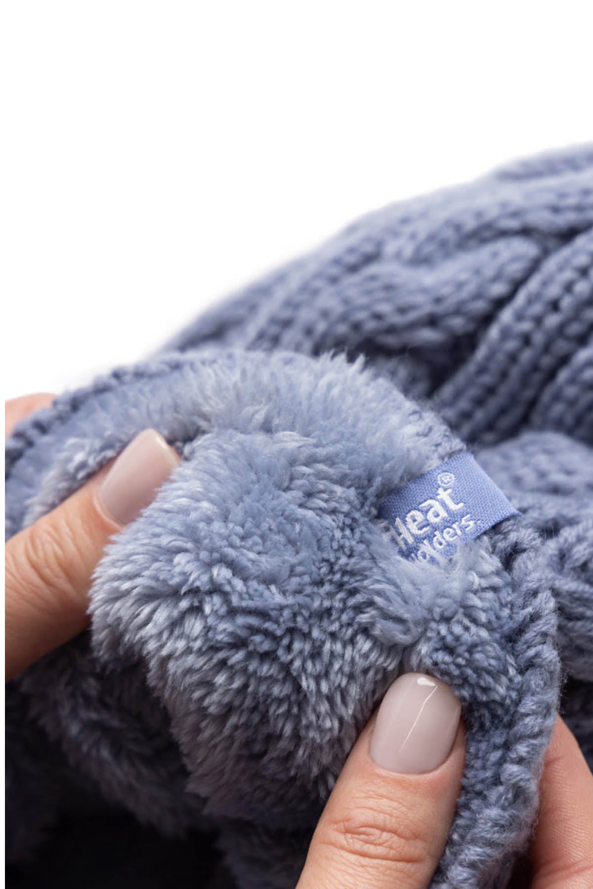 Close-up of hands holding a soft, light blue, fluffy knit Heat Holders Alesund Ladies Thermal Beanie with a small blue tag reading "Heat Holders." The beanie's texture appears warm and cozy, enhanced by its HeatWeaver® plush lining. The person's fingernails are neatly manicured with a light beige polish.