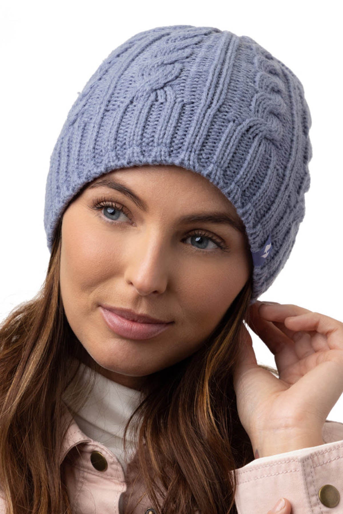 A woman with long brown hair is wearing a Heat Holders Alesund Ladies Thermal Beanie in light purple. She is seen touching the edge of the beanie with one hand. Dressed in a light-colored jacket, she looks slightly off to the side with a gentle expression against a white background.