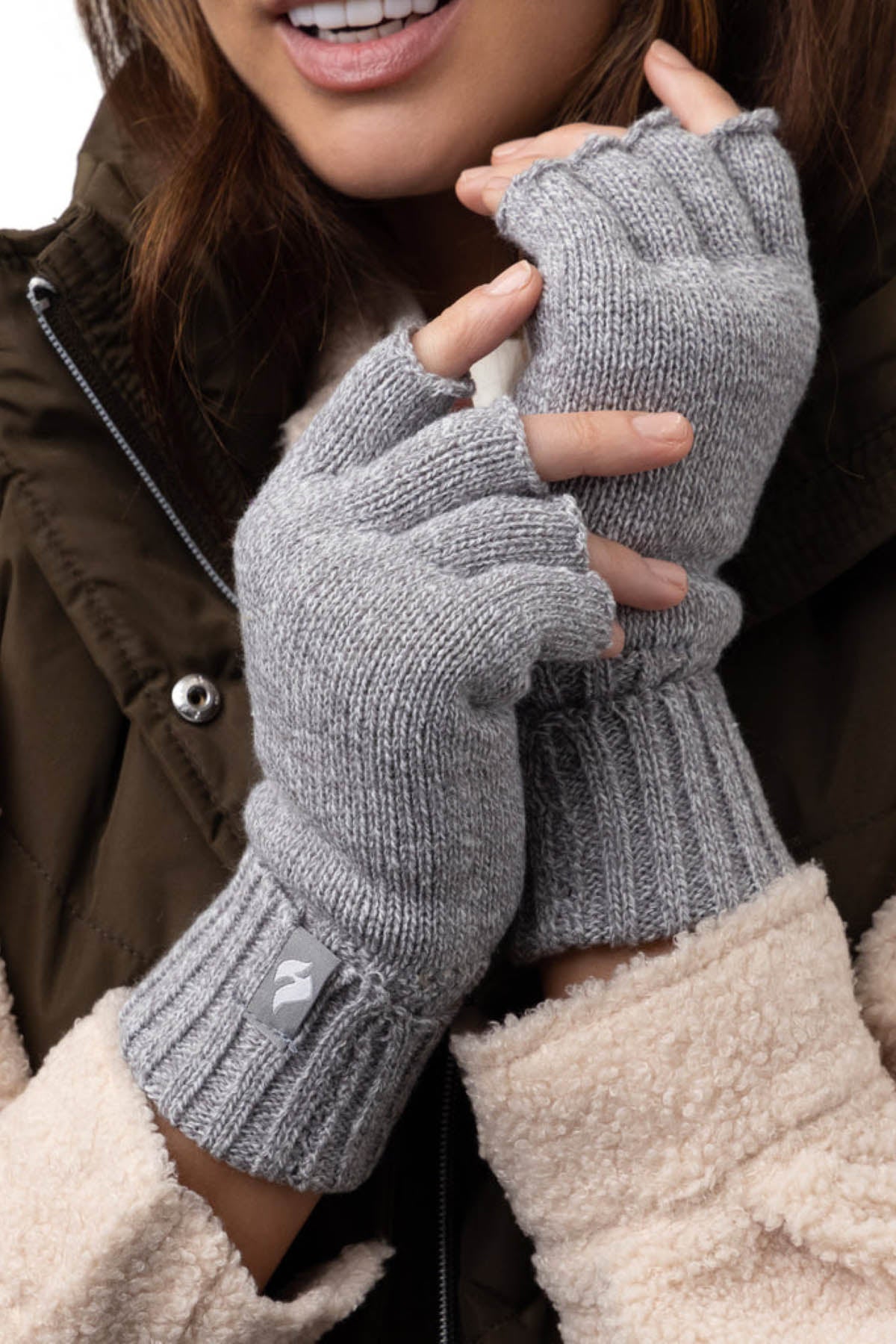Close-up of a person wearing Heat Holders Ladies Fingerless Thermal Gloves in gray and a brown jacket with cream-colored fleece sleeves. The person is holding their hands close together, partially overlapping, and smiling. Their hair is visible and they are outdoors in a casual setting.