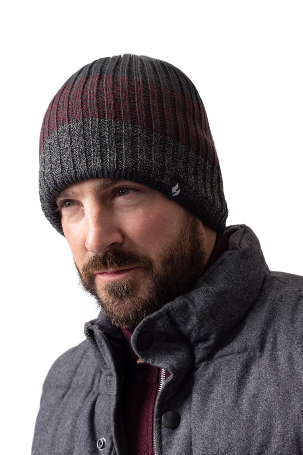 A bearded man wearing a Heat Holders Linden Block Stripe Men's Thermal Beanie in dark tones of gray and maroon, paired with a gray, quilted jacket featuring HeatWeaver lining. He gazes slightly to the left with a serious expression, standing in front of a plain white background, ready to face the cold weather.