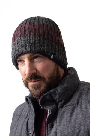 A bearded man wearing a Heat Holders Linden Block Stripe Men's Thermal Beanie in dark tones of gray and maroon, paired with a gray, quilted jacket featuring HeatWeaver lining. He gazes slightly to the left with a serious expression, standing in front of a plain white background, ready to face the cold weather.