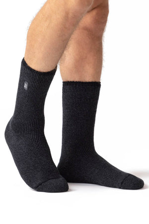 A person is wearing Heat Holders Mens Wool Sock—thick, black, ribbed mid-calf wool socks with a small rectangular label near the top—while standing against a white background, ideal for outdoor activities.
