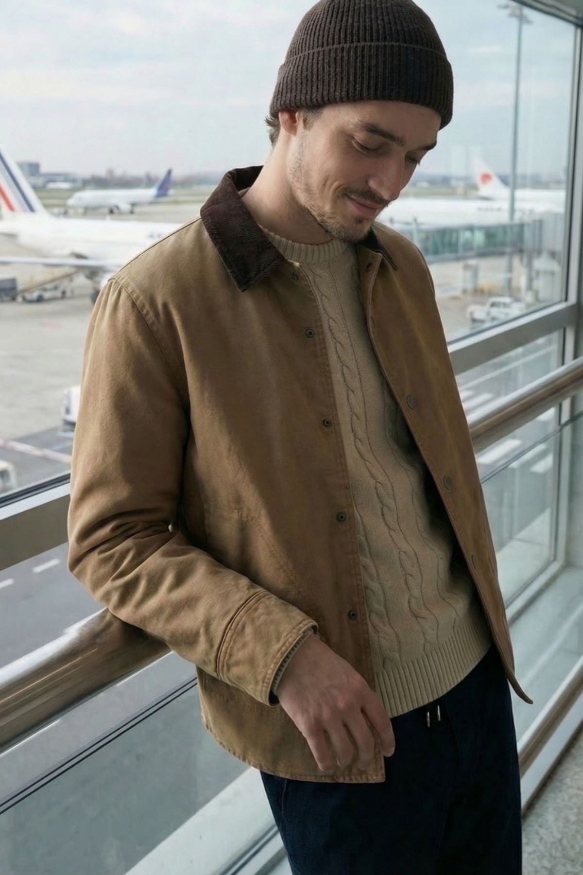 A man wearing a Hechter Patrick Canvas Jacket, beige sweater, and dark beanie stands by an airport window, smiling slightly as he looks down. Airplanes can be seen outside on the runway.