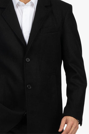 A person wears a black Howard Hudson Overcoat over a white dress shirt, pictured from neck to mid-thigh with their right arm slightly bent at the elbow.